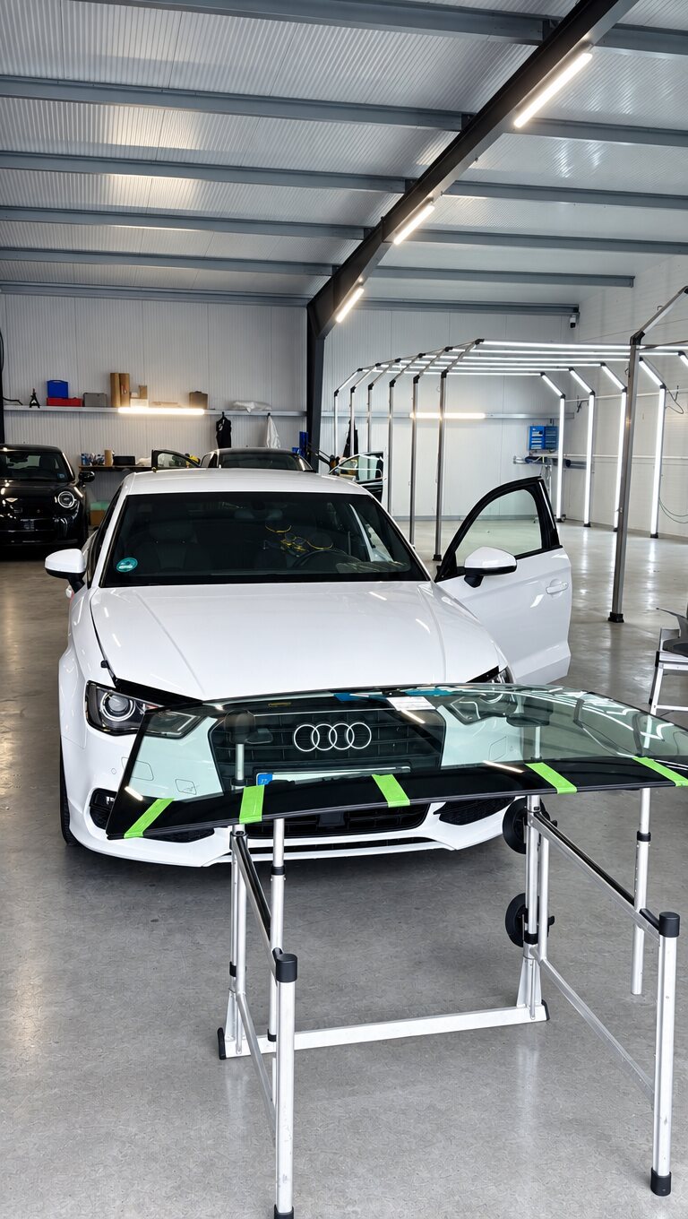Weißer Audi in der Halle, davor die abgeklebte neue Frontscheibe auf dem Montageständer